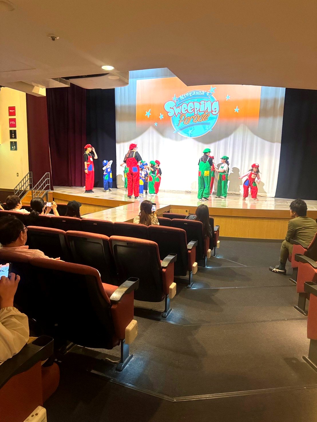キッザニア東京のシアター。ステージで子どもたちがカラフルな衣装でショーを上演し、客席では保護者が鑑賞している